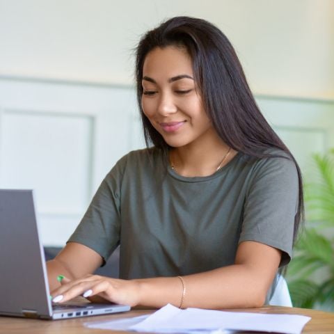 A woman works with papers at home in front of a laptop monitor. She is sitting with glasses and checking papers in front of laptop.