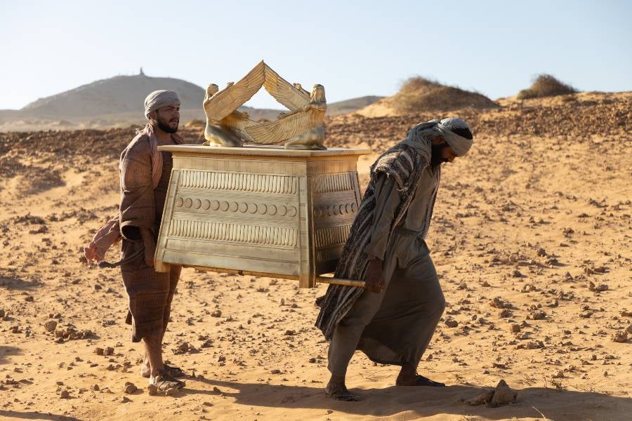 Two men carrying a replica of the Ark of the Covenant in a dessert in a scene from a movie. 