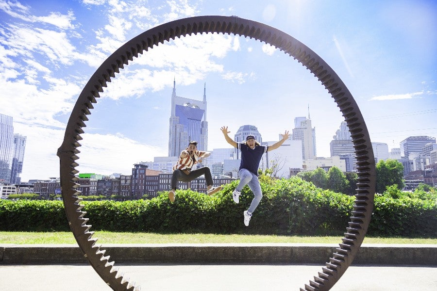 Austin & Colin jumping up into the air at Nashville's Riverfront Park, framed by a circular sculpture and the Nashville skyline in the background