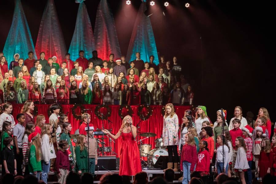 Ellie Holcomb singing on stage at the Lighting of the Green 2025 surrounded by choirs from Lipscomb Academy. 