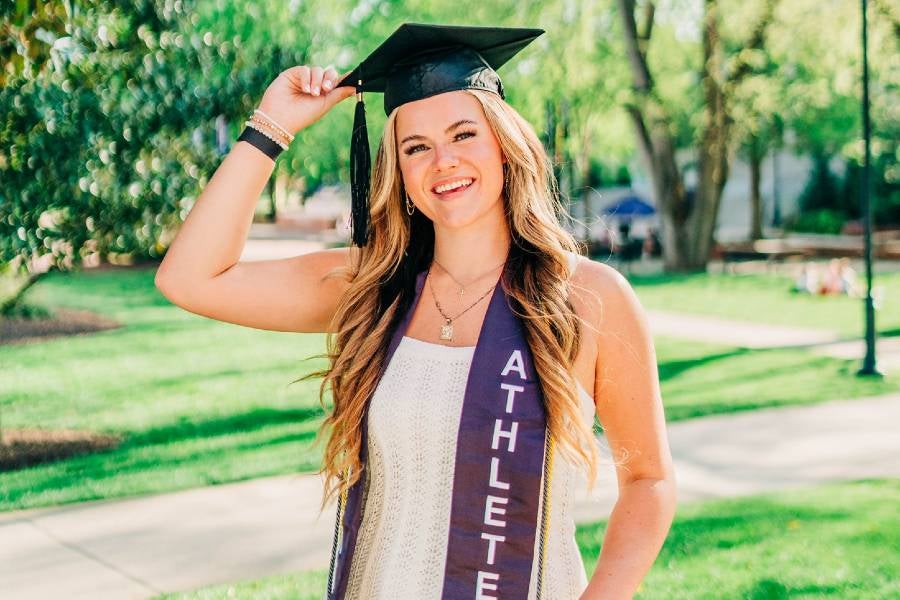 Meg Mersman standing in Bison square wearing a white dress, her athletics graduation shash and her graduation cap. 