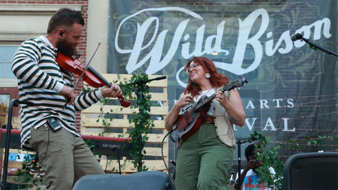 2 musicians playing guitar and fiddle in front of a Wild Bison Festival banner