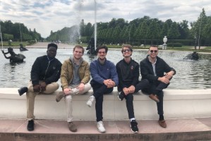 Five students sitting on a wall by a fountain. 