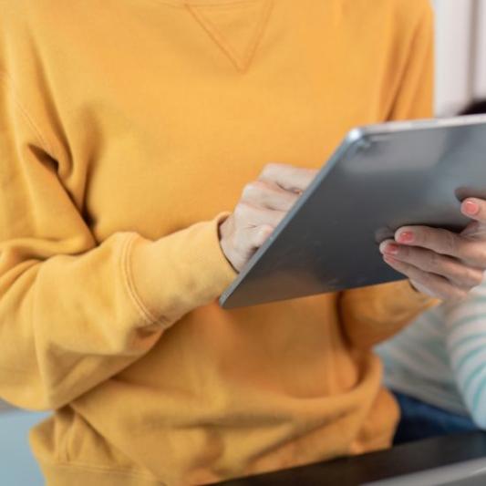 A close up of a mother on her tablet while her daughter does homework in the background.
