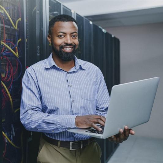 IT professional smiling while holding a laptop in a server room.