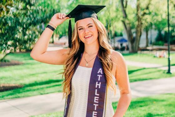 Meg Mersman standing in Bison square wearing a white dress, her athletics graduation shash and her graduation cap. 