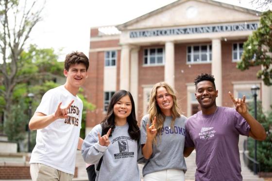 Four students standing in front of Collins Alumni Auditorium holding up "horn's up" signs. 